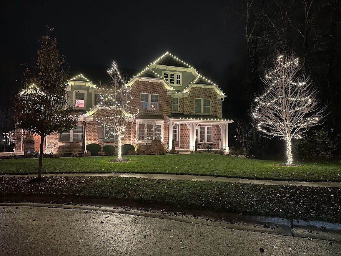 Brick two-story home with yard trees wrapped in warm white string lights